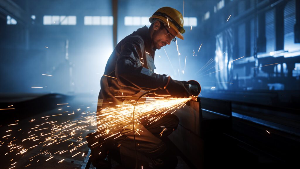 Industrial worker in safety gear using an angle grinder, creating bright sparks while cutting metal in a dimly lit factory.